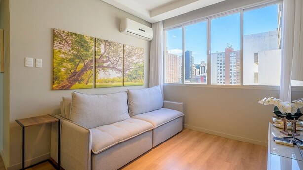 A modern living room with a beige sofa, a painting of trees, a window with a city view, and a small table with a vase of flowers. - Charming Apartment in Downtown Floripa Edc402 (Florianópolis)