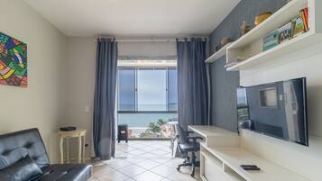 A modern living room with a large TV, a sofa, a small table, and a view of the sea through large windows.