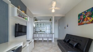 A modern living room with a black leather sofa, a flat-screen TV mounted on the wall, and a kitchen area with white cabinets and a tiled backsplash. - Charming Apt With Balcony and Sea View Edj0410 (Florianópolis)