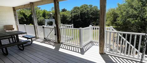 A covered outdoor deck with a picnic table and chairs, white railings, and a view of trees.