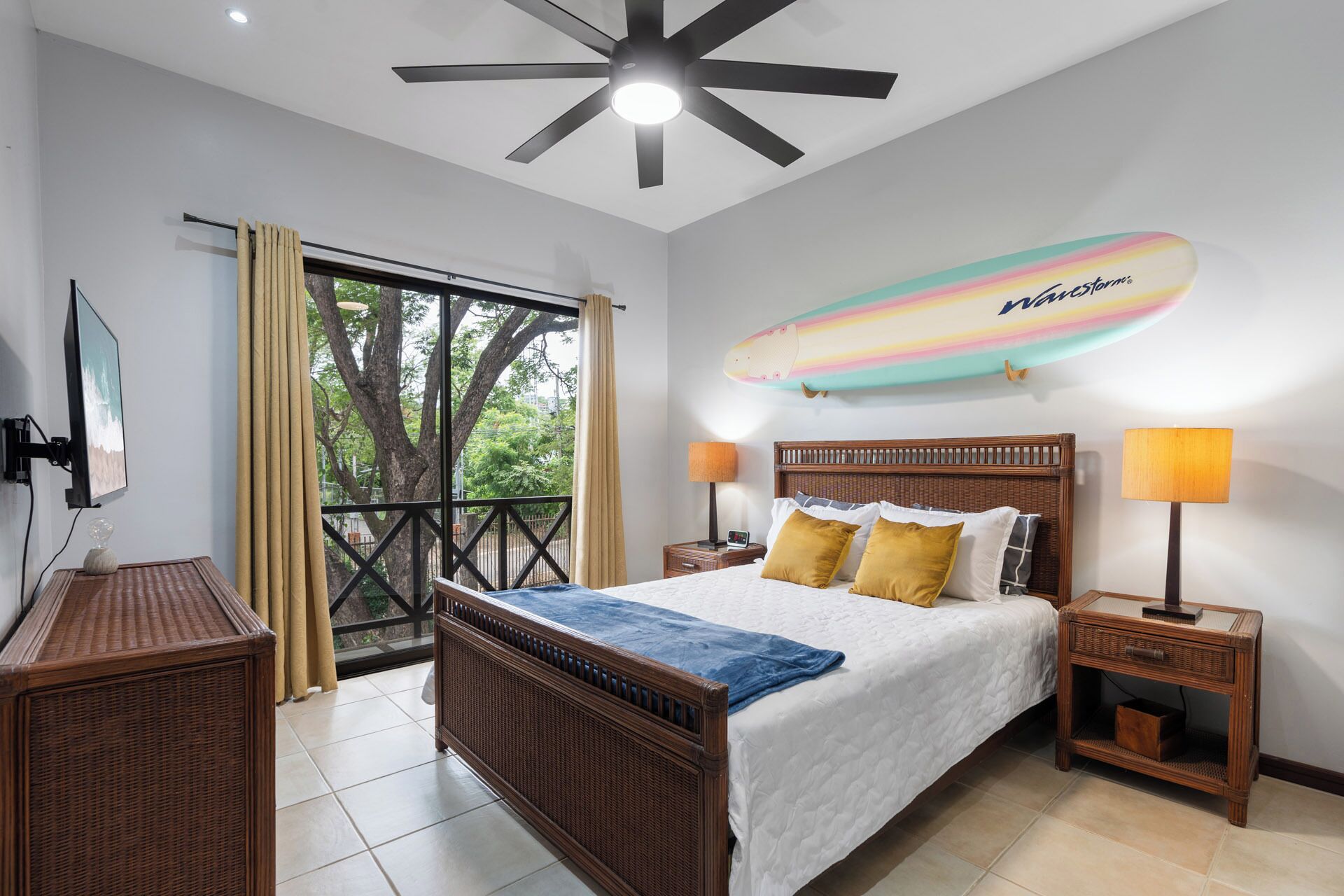 A bedroom with a bed, nightstands, a ceiling fan, a beach-themed surfboard decoration, and a view of trees through a large window.