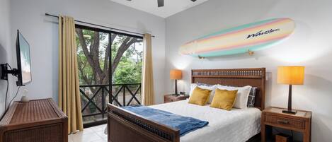 A bedroom with a bed, nightstands, a ceiling fan, a beach-themed surfboard decoration, and a view of trees through a large window.