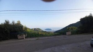 Une route asphaltée, bordée d’une clôture en métal et de verdure d’un côté, qui mène à un paysage vallonné offrant une vue lointaine sur la mer.