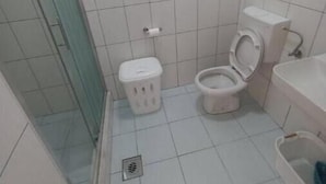 A small bathroom with white tiles, a toilet, a trash bin, and a shower area.