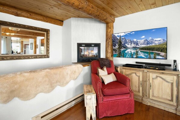 A cozy living room with a fireplace, a red checkered armchair, a large flat-screen TV, and a wooden ceiling.