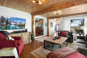 A spacious living room with a wooden ceiling, a large flat-screen TV, a fireplace, and a view of the mountains through the windows.