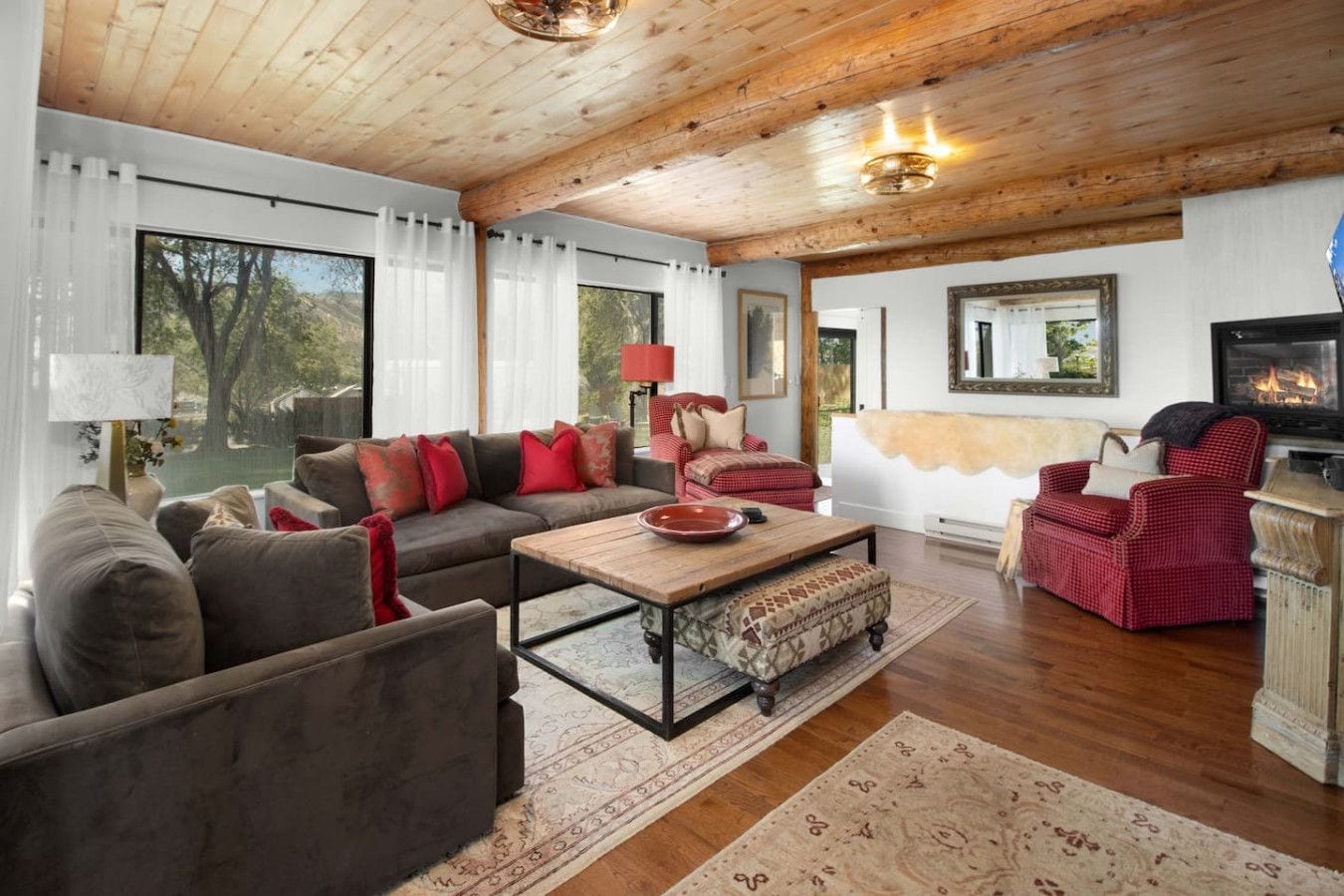 A cozy living room with a wooden ceiling, a fireplace, and a large window with curtains.