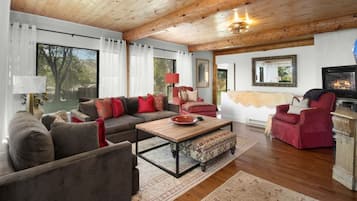 A cozy living room with a wooden ceiling, a fireplace, and a large window with curtains.