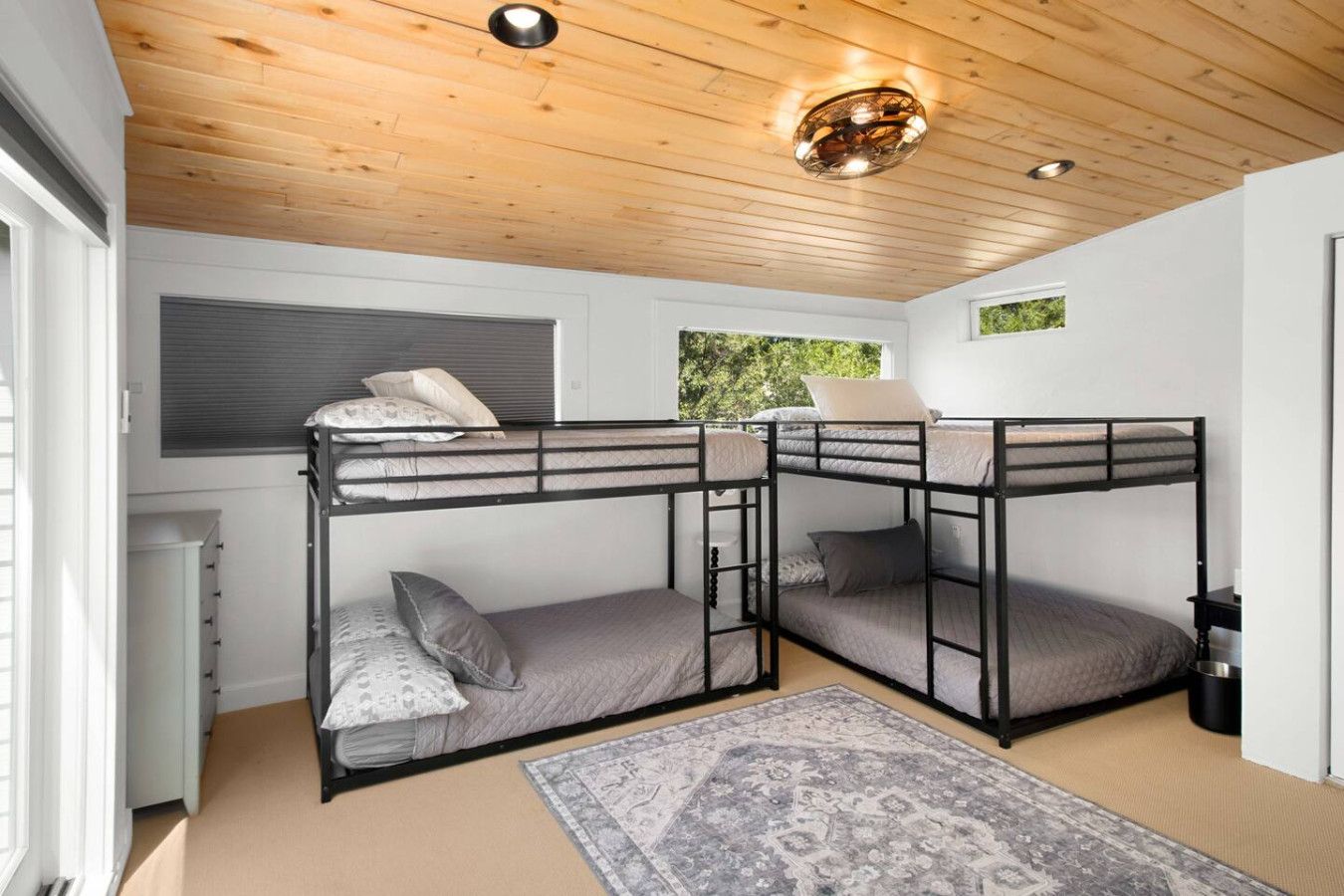 Une chambre avec des lits superposés, un plafond en bois, une fenêtre donnant sur un paysage verdoyant et un tapis à motifs sur le sol.