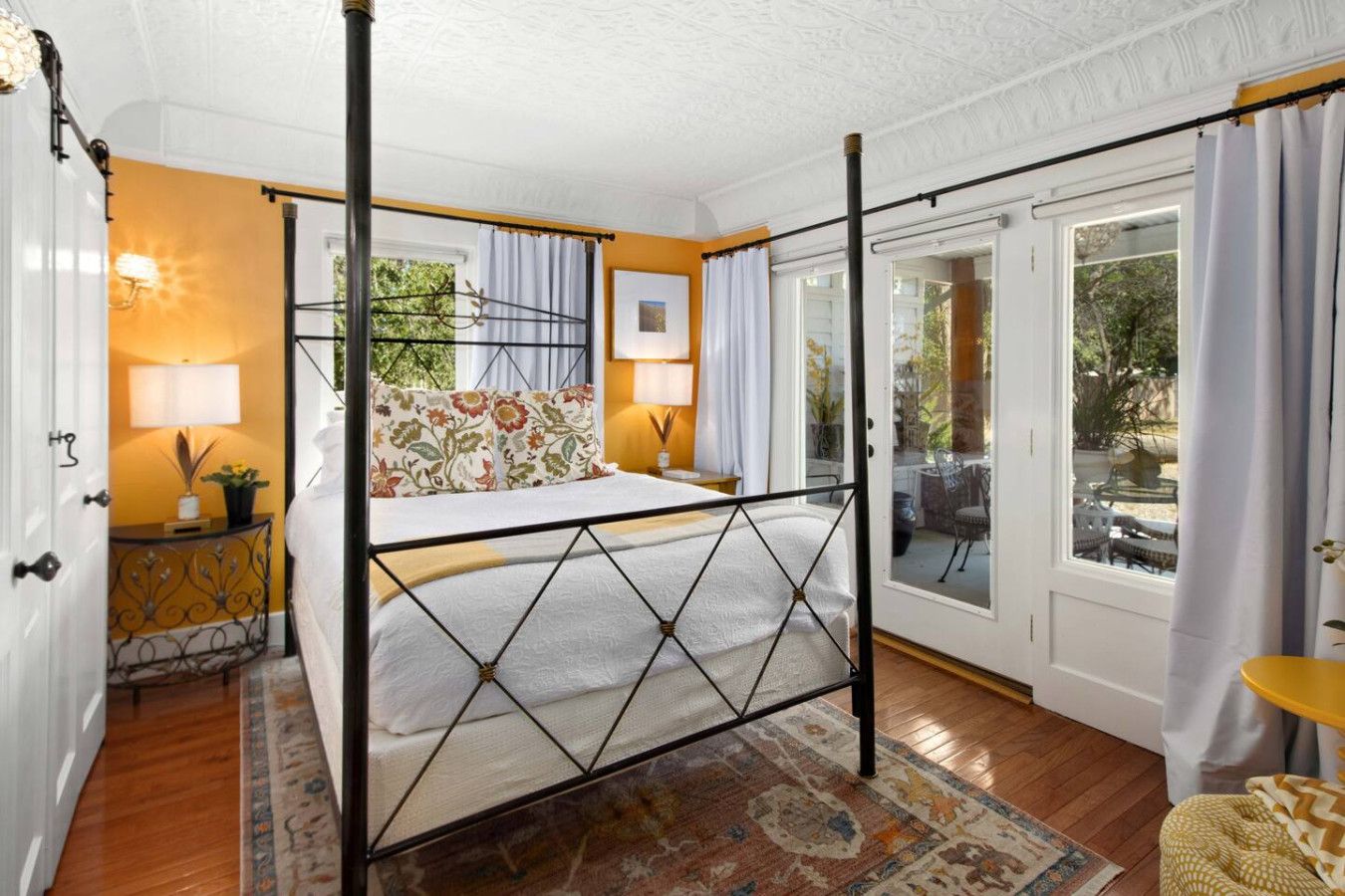 A bedroom with a four-poster bed, a nightstand with a lamp, a window with curtains, and a view of the outdoors.