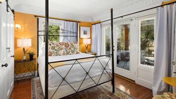 A bedroom with a four-poster bed, a nightstand with a lamp, a window with curtains, and a view of the outdoors.