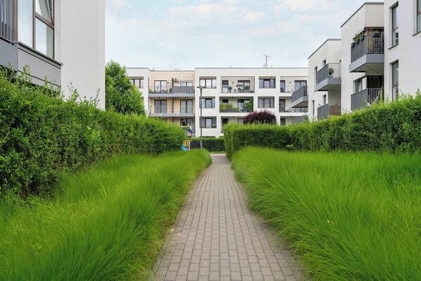 Un camino bordeado por setos verdes y frondosos que conduce a modernos edificios de apartamentos con balcones y plantas de exterior.