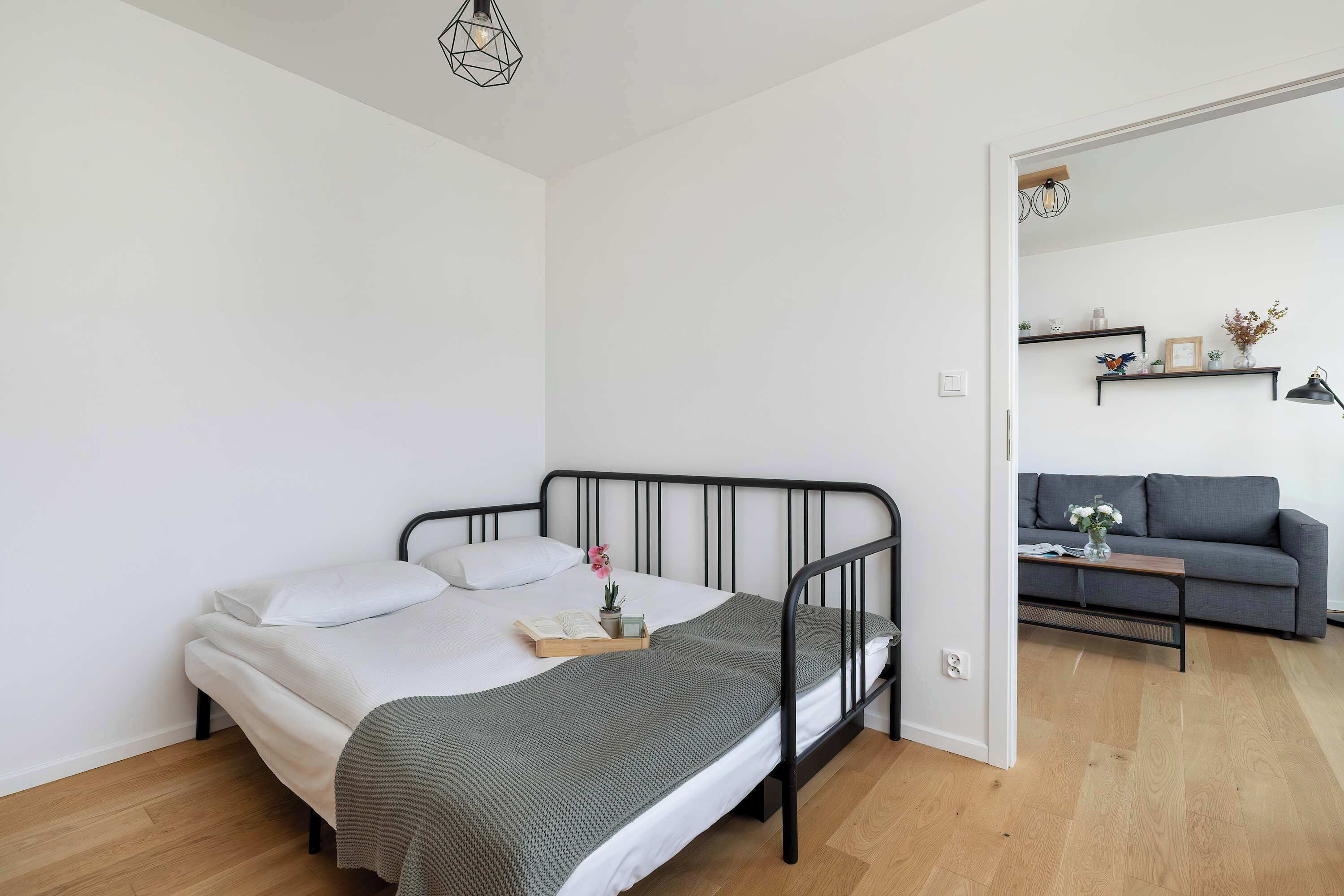A bedroom with a bed, bedside table, and a small desk with a chair. There is a sofa and a coffee table in the adjacent living area.