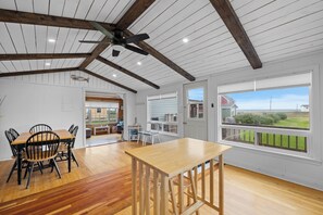 Una habitación amplia con una mesa y sillas de madera, un ventilador de techo y ventanas grandes que ofrecen vistas al exterior.