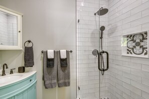 A bathroom with a white tiled shower, a sink with a mirror, and a vanity with a blue cabinet. - Your Breezy Old Town Retreat Awaits (Key West)