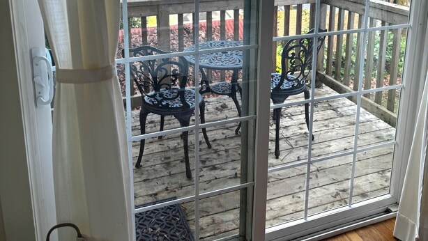 Une porte-fenêtre coulissante donne sur un patio extérieur doté d’une terrasse en bois et de chaises en métal.