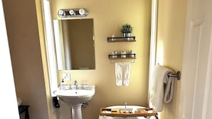 Une salle de bain avec un lavabo sur pied blanc, une baignoire îlot et un miroir au-dessus du lavabo. Il y a deux étagères au-dessus de la baignoire, l’une servant de support à une plante et l’autre à des articles de toilette.