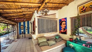 Um quarto com um sofá trançado, um banquinho verde, um espelho decorativo e um teto com vigas de madeira.