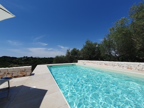 A clear blue swimming pool with stone walls, surrounded by a tiled deck and trees. - Trullo Antonelli (Puglia)