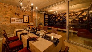 Une salle à manger avec une longue table dressée pour un repas, une cave à vin visible à travers une cloison vitrée et un mur en briques.