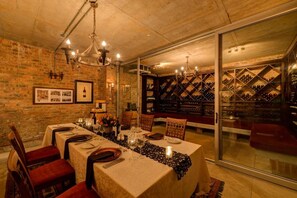 Une salle à manger avec une longue table dressée pour un repas, une cave à vin visible à travers une cloison vitrée et un mur en briques.