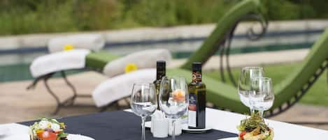 A dining table with plates of food, wine glasses, and bottles, set outdoors with a pool and greenery in the background.
