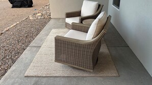 Una sedia in vimini con un cuscino su un tappeto, un barbecue coperto e un patio in cemento con una recinzione sullo sfondo.
