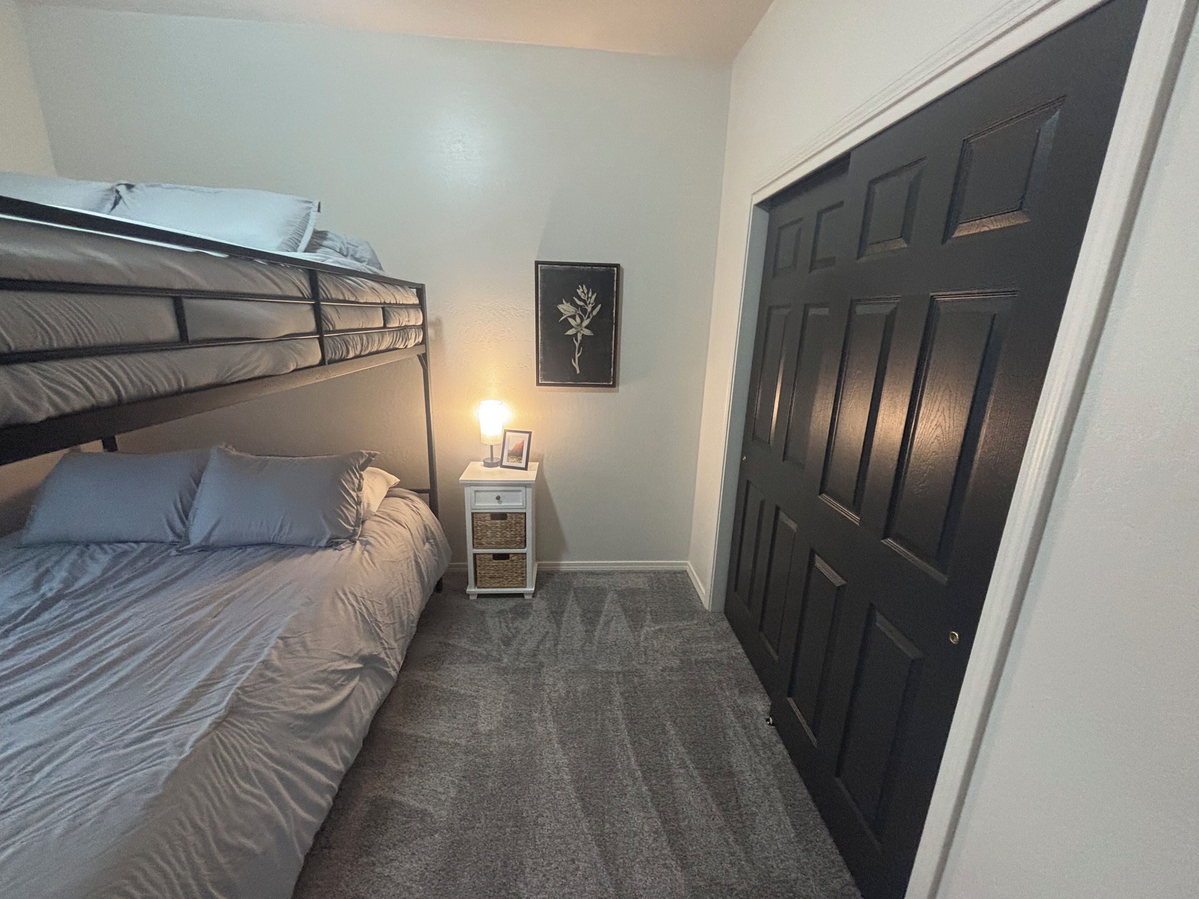 Quarto com cama beliche, mesa de cabeceira com abajur, quadro na parede e porta escura.
