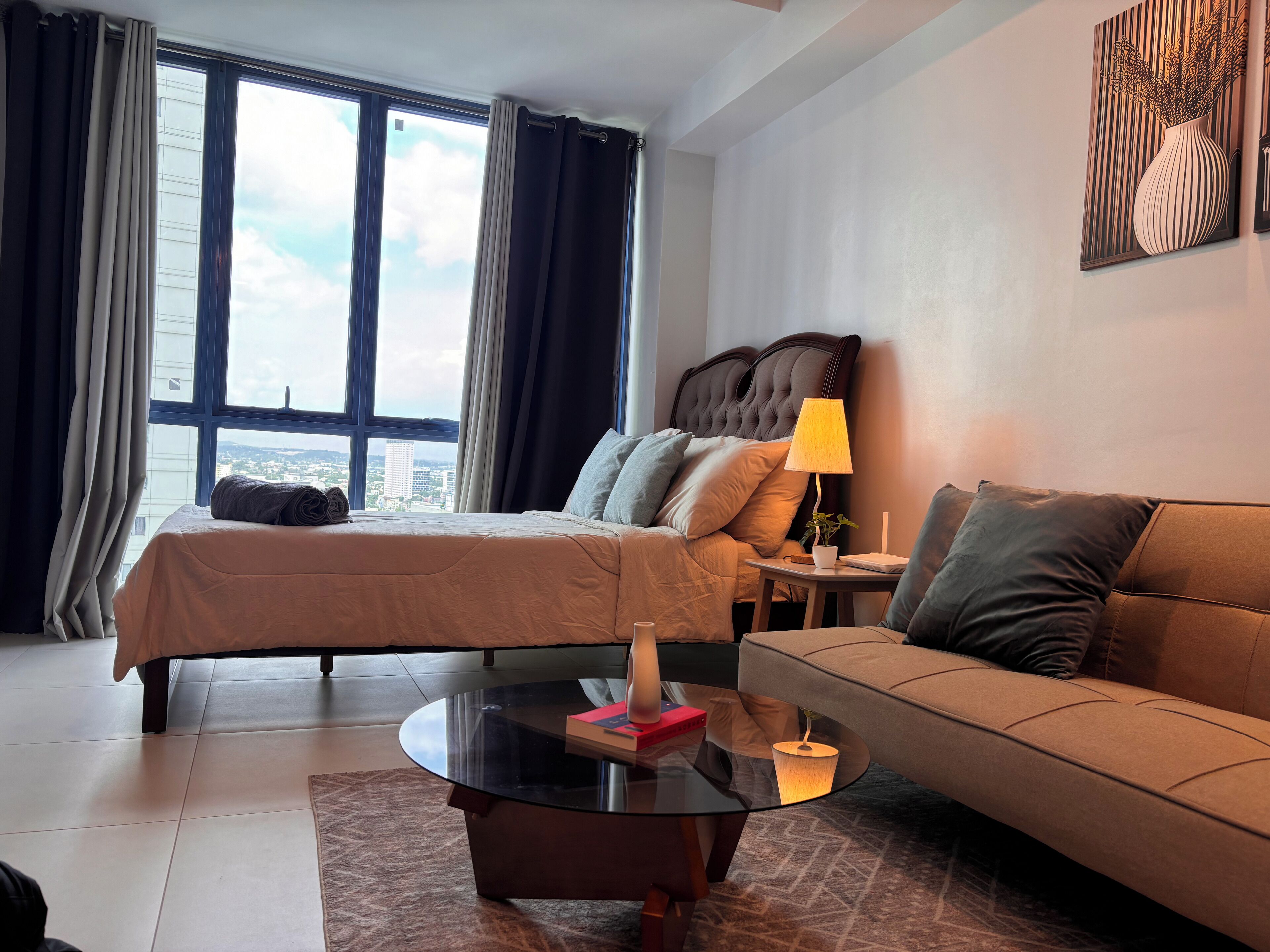 Habitación de hotel con cama, sofá, una mesita de centro de vidrio y vistas a la ciudad a través de amplias ventanas.