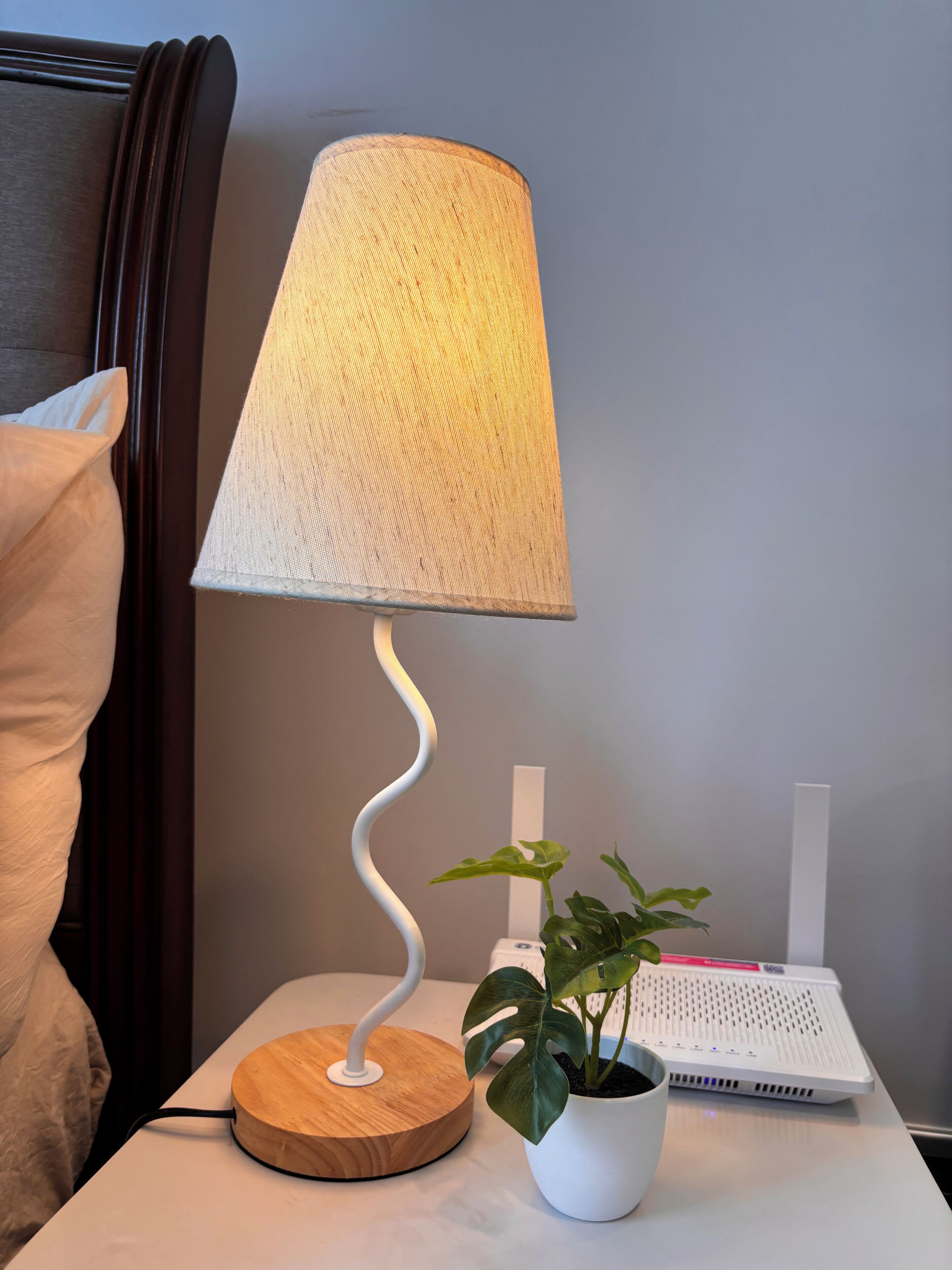 Una mesita de noche con una lámpara, una planta en maceta y un dispositivo electrónico blanco.