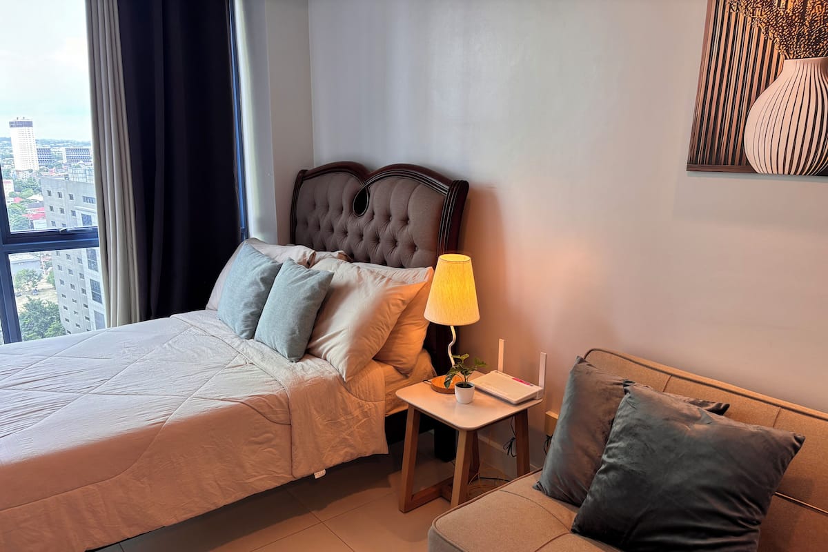 Habitación de hotel con una cama grande, una mesita de noche con lámpara, una pequeña mesa con una planta y un sofá con cojines.