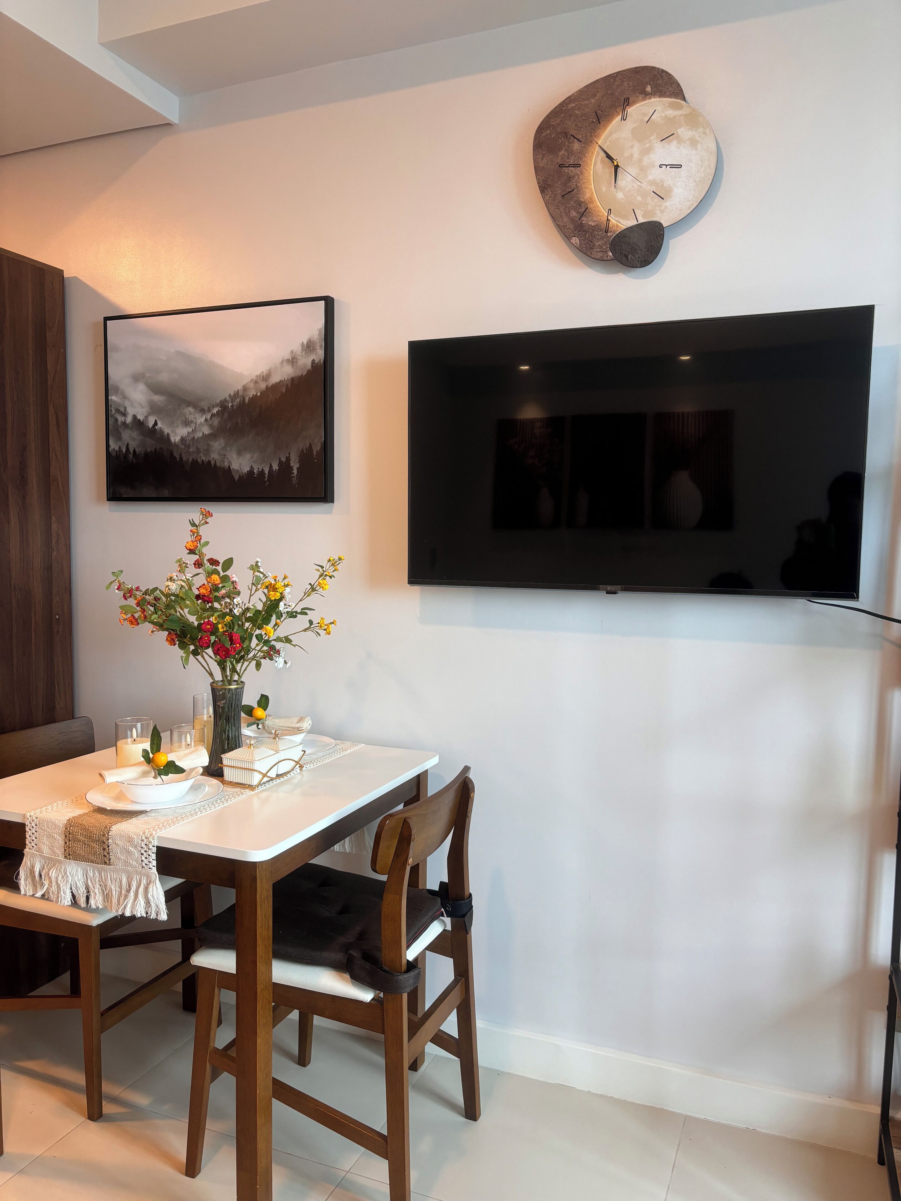 A dining area with a table set for two, a mounted TV, and a wall clock.