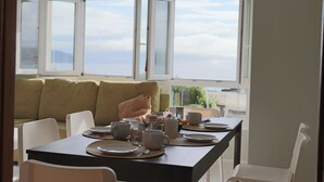 A dining area with a table set for two, a sofa, and a view of the sea through open windows. - Apartment in front of the beach of Coira in the center of Portosin (Goyanes)