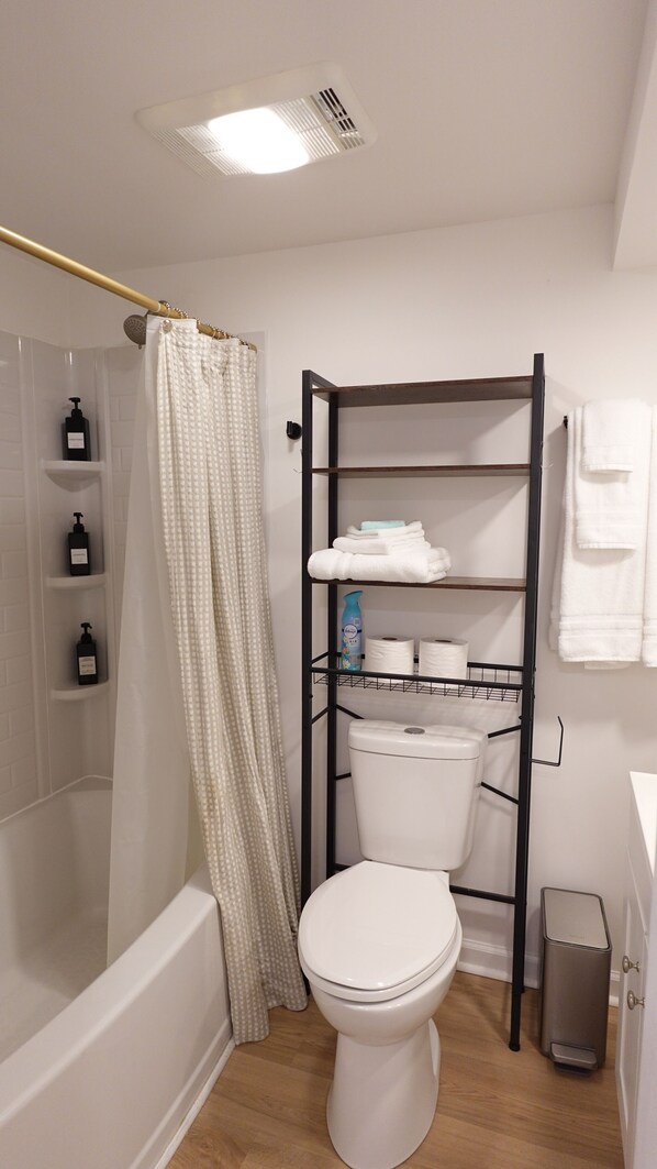 A bathroom with a white toilet, a shower with a beige curtain, a black metal shelf unit with towels, and a trash bin. - Cozy 3BR Retreat w/ Patio, Fire Pit & Yard (Winchester)