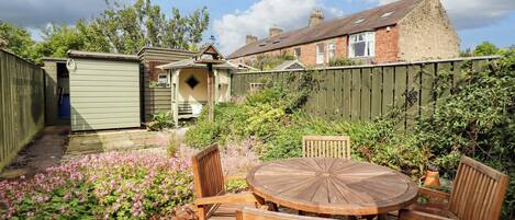 A patio with a round wooden table and chairs, surrounded by a garden with pink flowers and a brick wall.