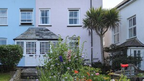 A two-story building with a light blue and white facade, a stone roof, and a garden with various flowers and a palm tree. - 84 Fore Street (Port Isaac)