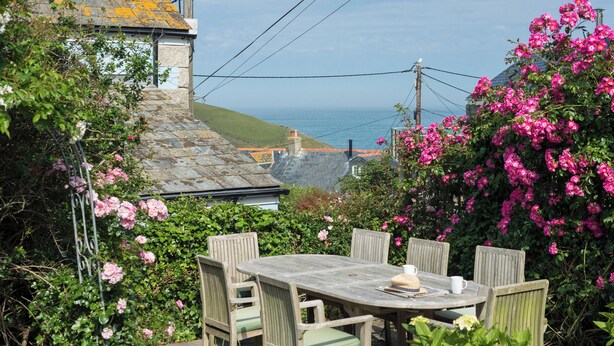 A round wooden table with four chairs, surrounded by lush greenery and vibrant pink flowers. - 84 Fore Street (Port Isaac)