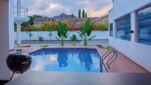 A modern outdoor pool area with a barbecue grill, surrounded by lush greenery and a clear view of the neighborhood.