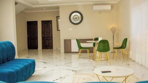 A modern living room with a blue sofa set, a round coffee table, and a dining area with green chairs.