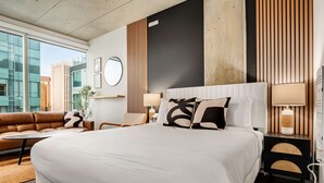 Une chambre d’hôtel moderne avec un grand lit, un canapé, un miroir rond et une vue sur le paysage urbain.