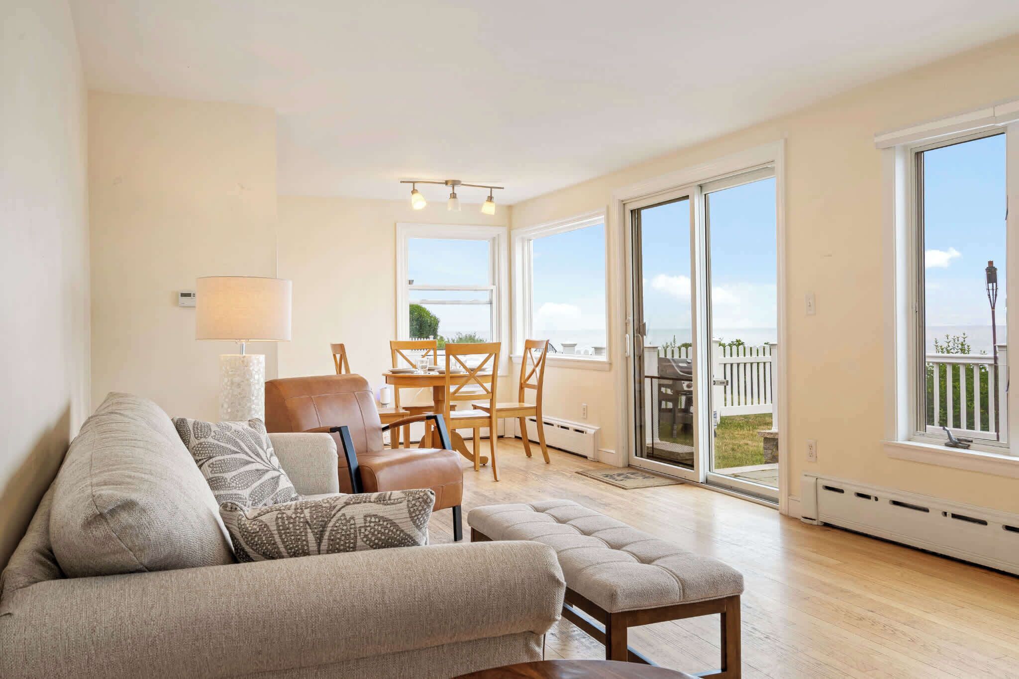 A living room with a sofa, ottoman, and a dining area with a table and chairs. There is a large window with a view of the outdoors.