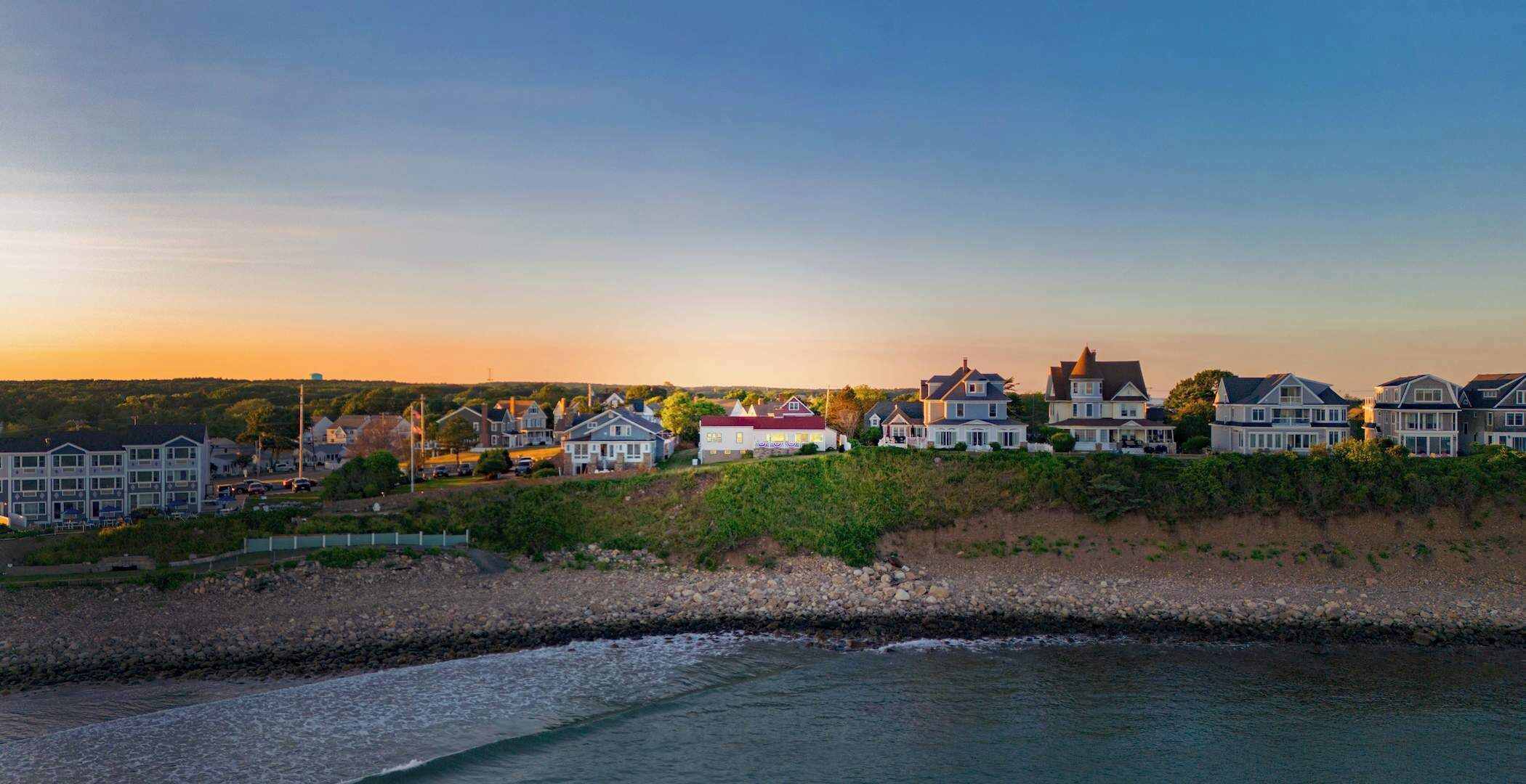 A coastal residential area with houses along a shoreline, featuring a mix of single and multi-story buildings, some with visible balconies and a variety of roof styles.