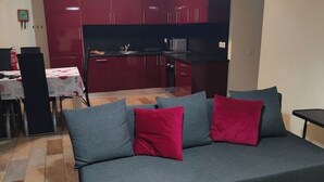 Ein Wohnzimmer mit grauem Sofa und roten Kissen, einem kleinen Tisch mit weißem Tuch und einer Küchenzeile mit roten Schränken und einer Mikrowelle.