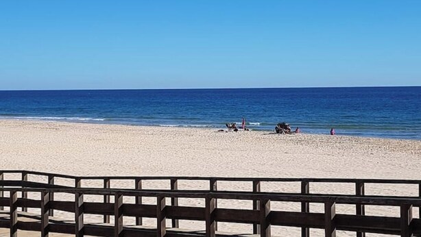Ein Strand mit einer Holzzahlung im Vordergrund und einem wolkenlosen, blauen Himmel.