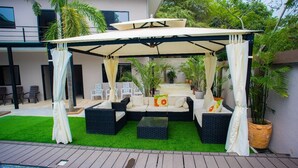 Un espace extérieur aménagé avec des sièges, un auvent, de l’herbe artificielle verte et des plantes en pot.