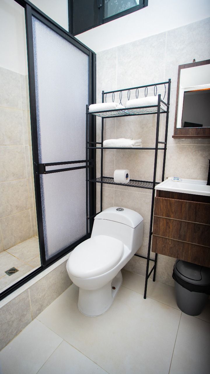 A modern bathroom with a white toilet, a glass shower enclosure, and a metal shelf unit with towels and toilet paper.
