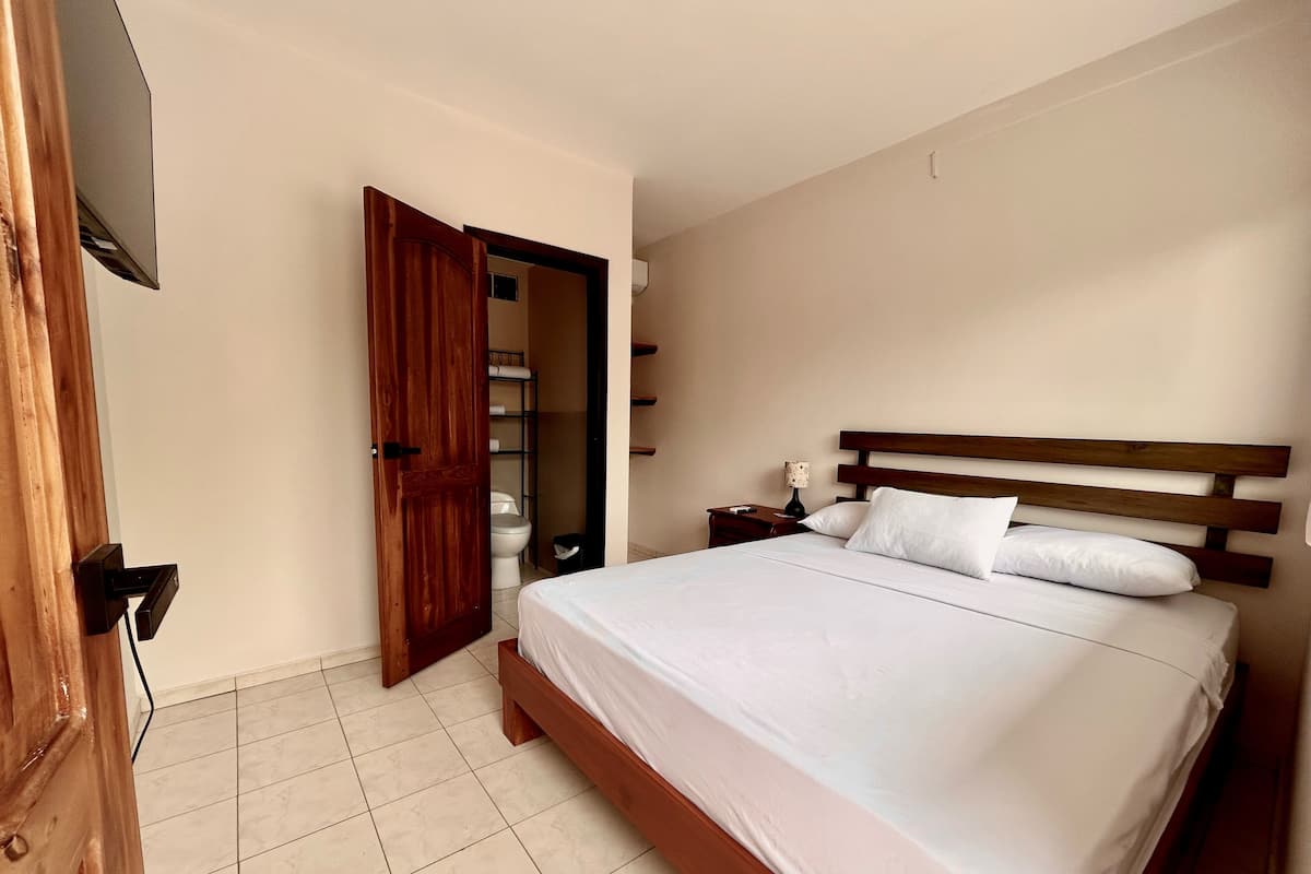 Ein Schlafzimmer mit einem hölzernen Bett, einem Nachttisch, einem an der Wand montierten Fernseher und einem durchgehenden Türspalt einsehbaren Badezimmer.
