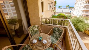A balcony with wicker furniture, a table with cups and saucers, and a view of the sea and buildings.