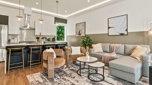 A modern living room with a grey sofa, two armchairs, a coffee table, and a bar area with stools. - Upscale 3BR | Sleeps 11 | Rooftop + Parking (Dallas)