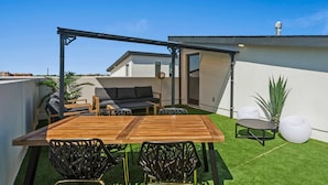 A rooftop patio with a wooden table, black chairs, a grey sofa, and a small round table with a white chair. - Upscale 3BR | Sleeps 11 | Rooftop + Parking (Dallas)